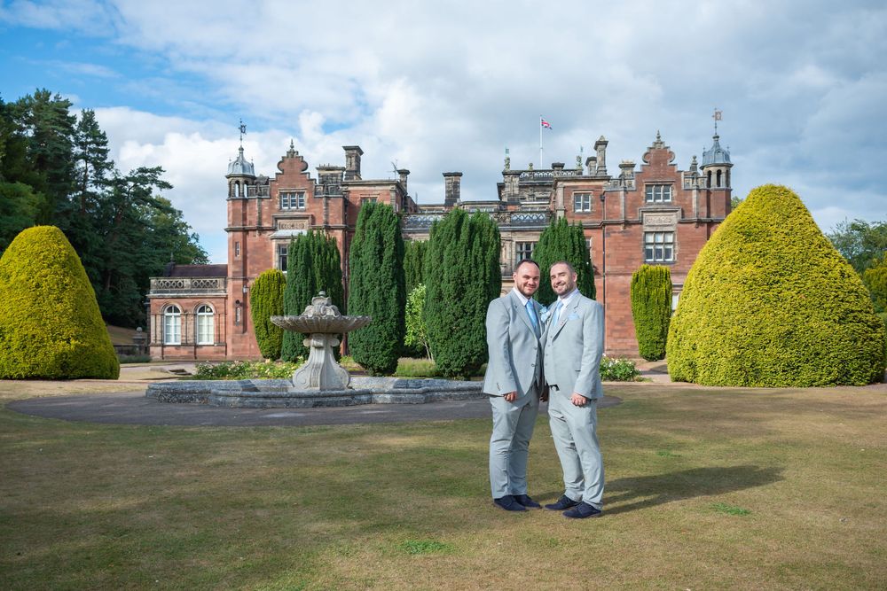 Two men in matching gray suits pose together on manicured grounds before a grand red brick Scottish manor house.