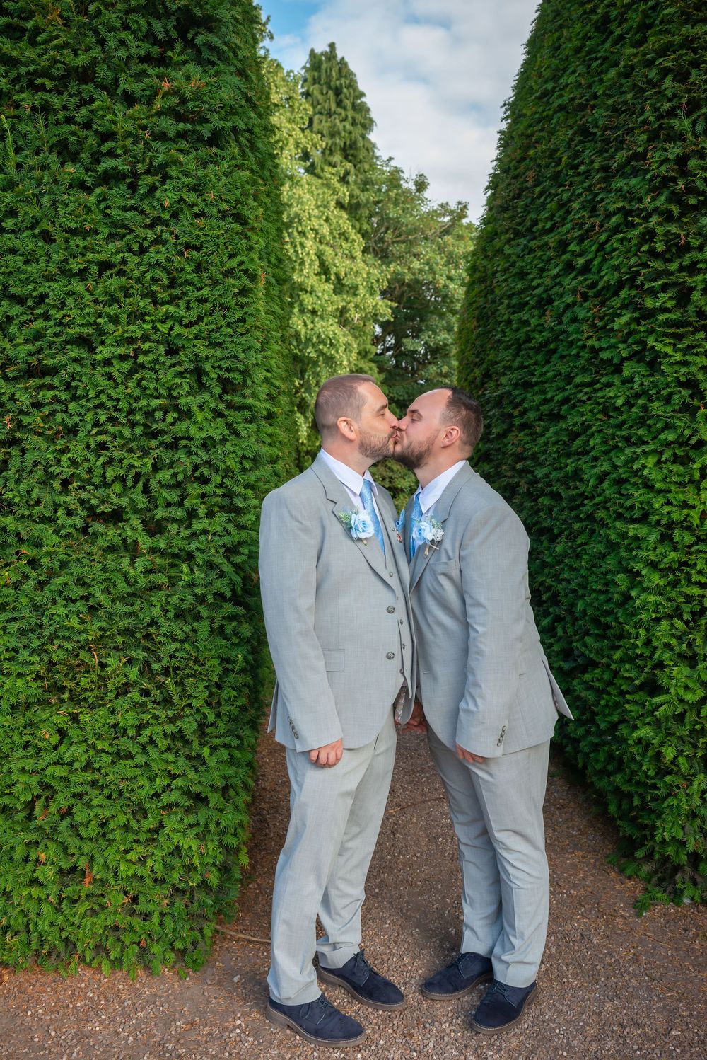 Two grooms in matching grey suits share a kiss in a lush green hedge garden pathway on their wedding day.