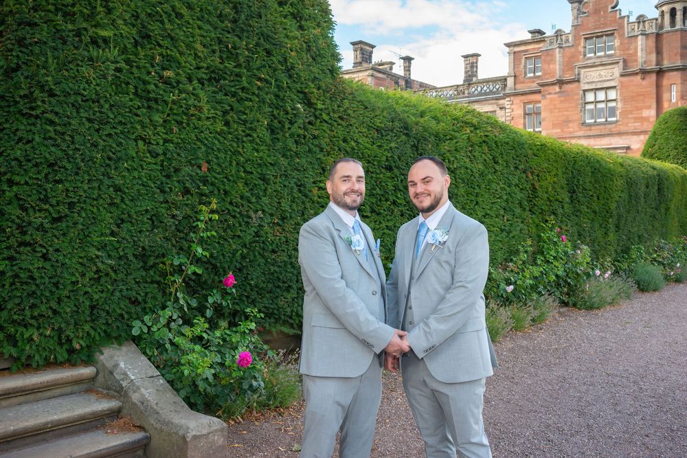 Two grooms in matching grey suits hold hands smiling outside a grand brick manor surrounded by tall hedges.