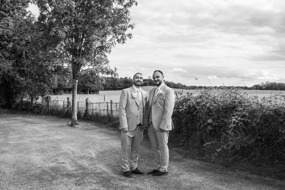 Two grooms in matching suits pose on a country path surrounded by trees and fields on their wedding day.