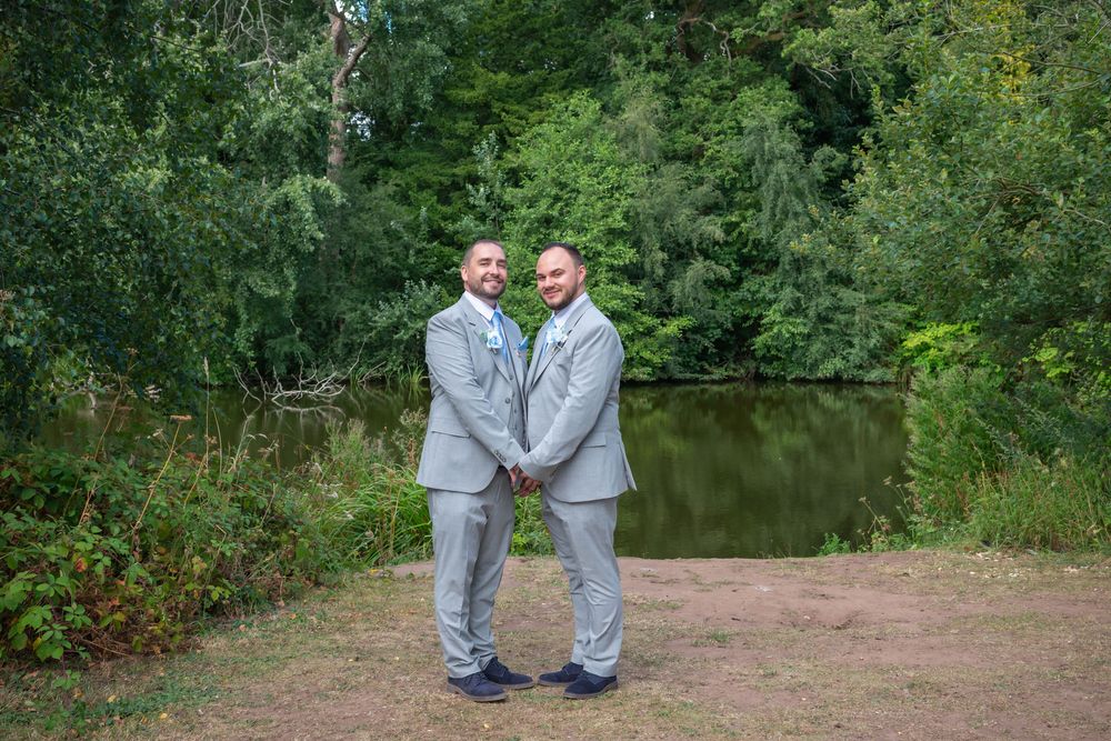 Two grooms in matching gray suits hold hands outdoors near a pond surrounded by lush green trees on their wedding day.