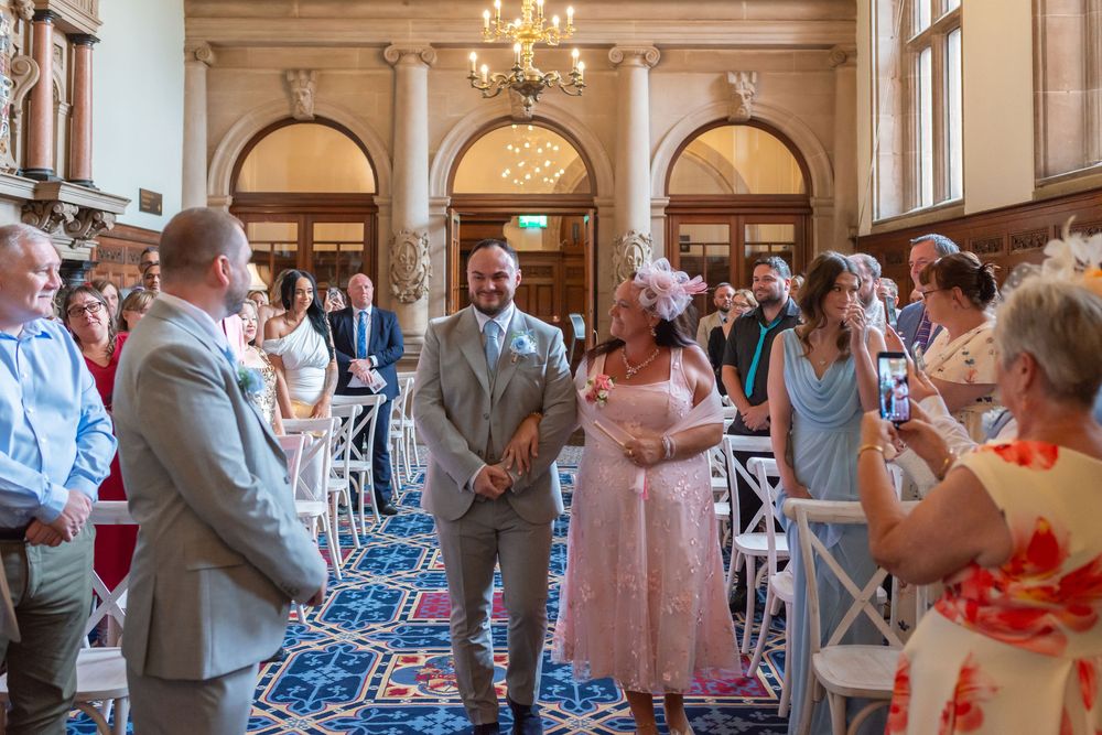 Groom walks bride down the aisle in an elegant chandelier-lit wedding venue with guests standing in celebration.