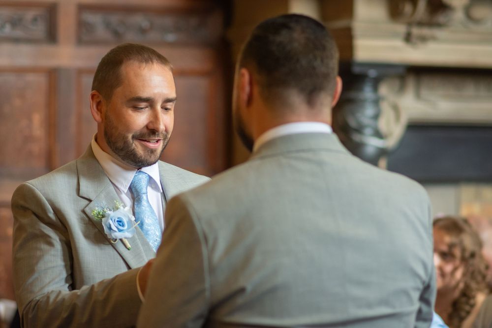 Two grooms in tan suits share a tender moment at their wedding ceremony near a stone fireplace.