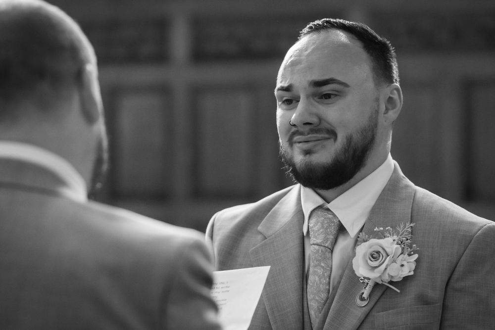 Groom in a beige suit with boutonniere smiling during wedding ceremony vows, black and white photo.