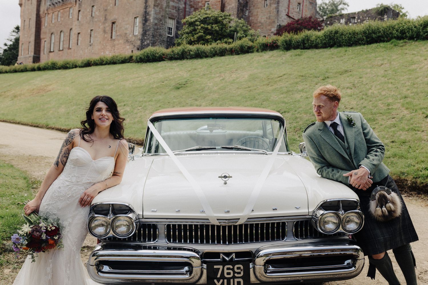 Bride and groom leaning against a vintage white car outside a Scottish castle wedding venue.