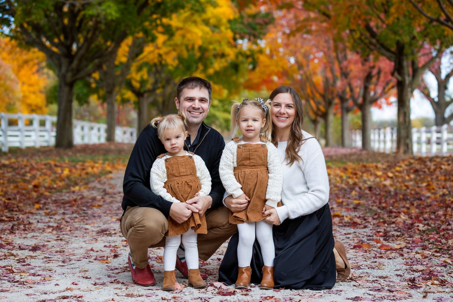 Family of three posing among autumn trees with colorful fall foliage, parents holding smiling baby outdoors.