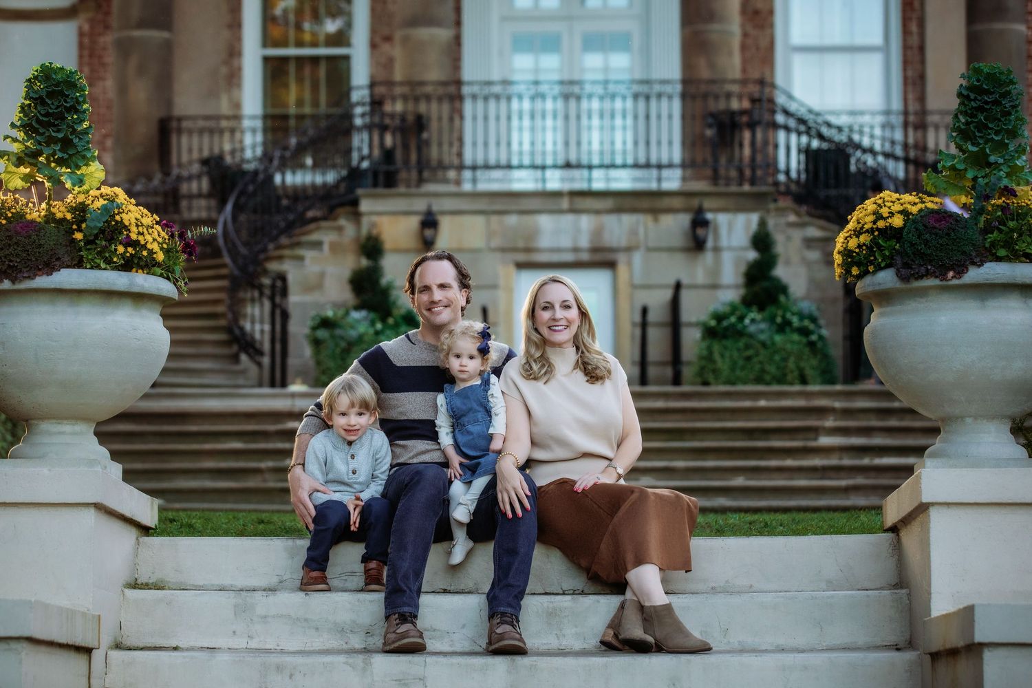 Family of three posing among autumn trees with colorful fall foliage, parents holding smiling baby outdoors.
