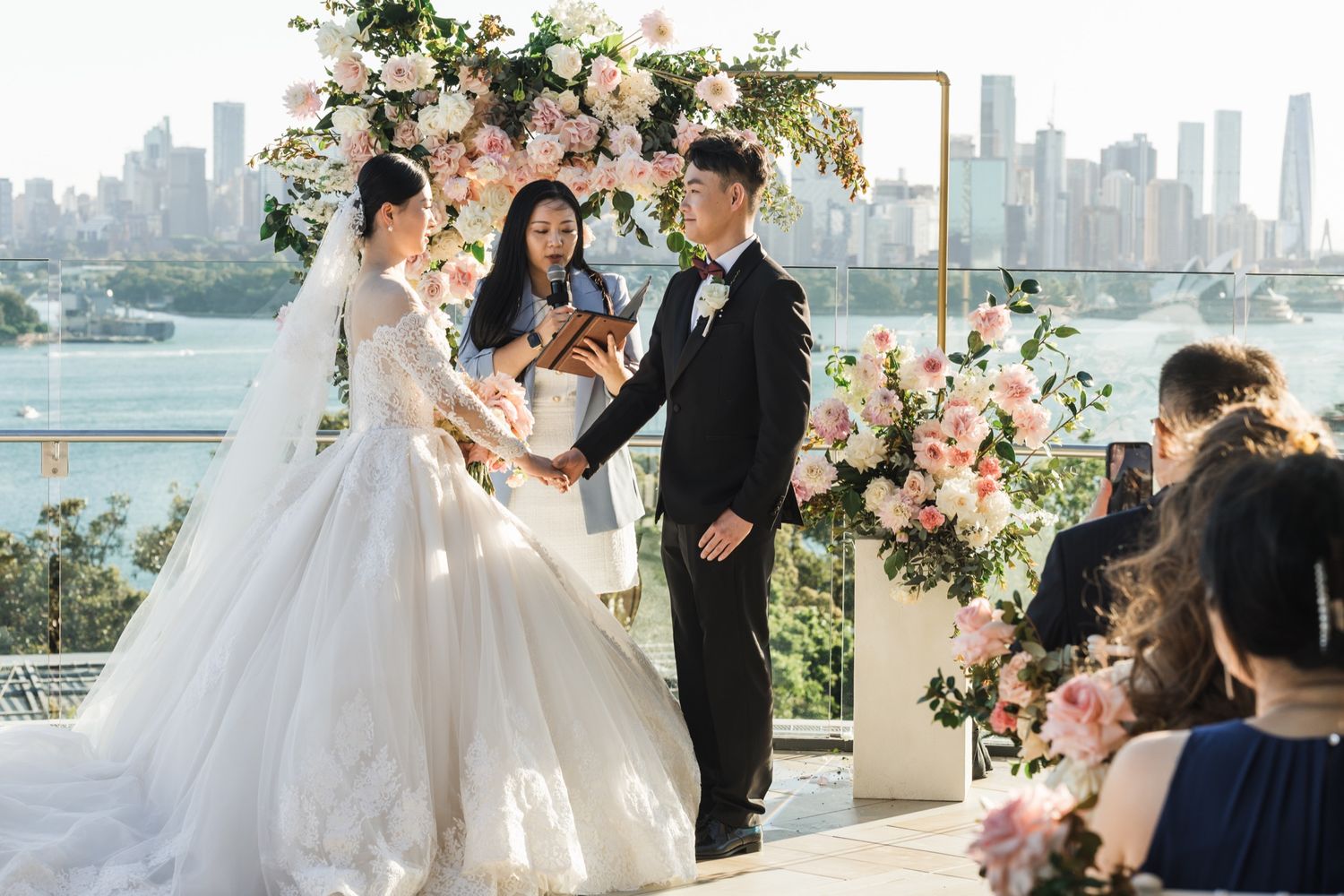 Couple exchanges vows at outdoor wedding ceremony with pink floral arch and city skyline backdrop.