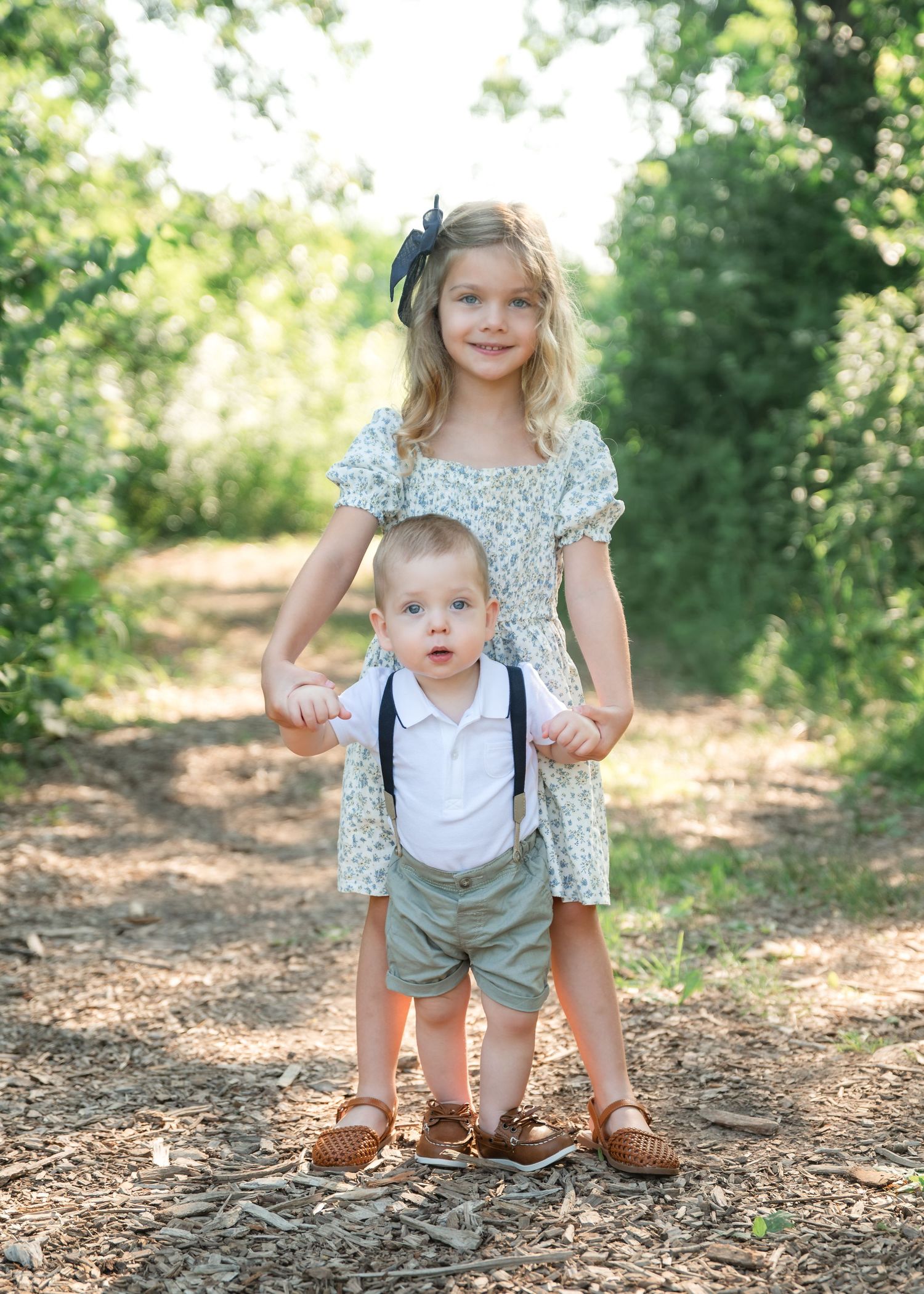 A young girl in a floral dress holds hands with her toddler brother in suspenders on a sunny outdoor path.