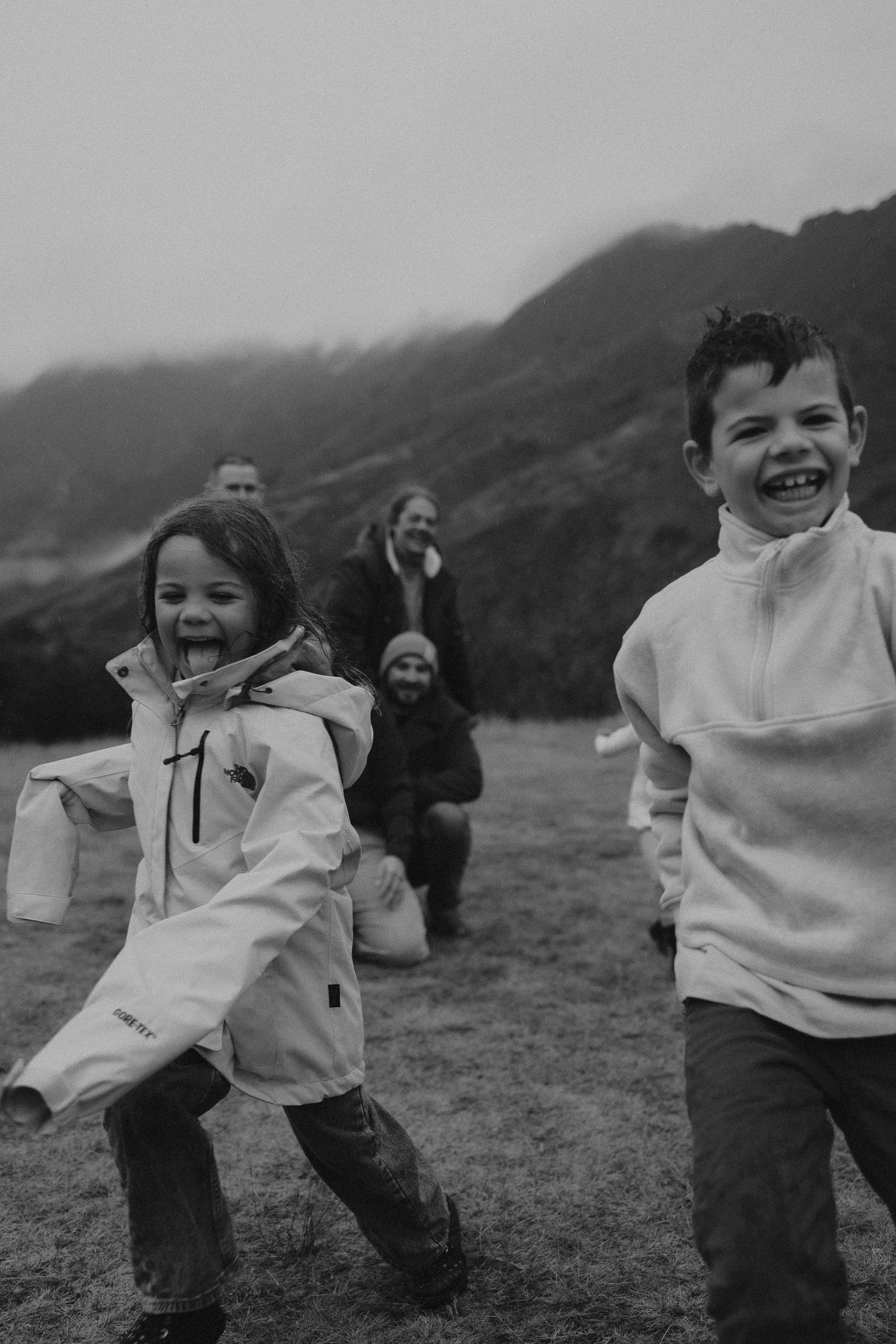 A cheerful toddler girl in a white sweater and blue pants runs along a lakeside with mountains in the background.
