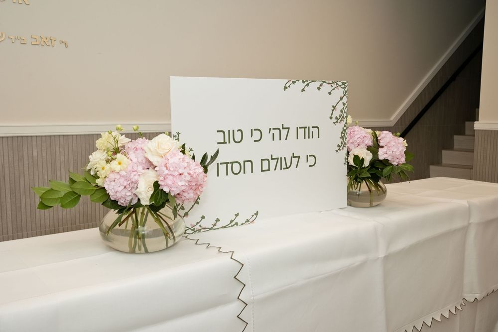 Hebrew sign on white tablecloth with pink and white floral arrangements at a Jewish celebration event.