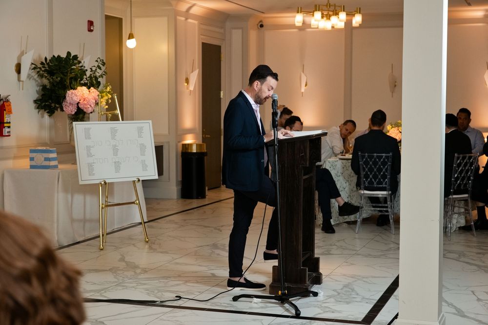 Man in suit speaking at podium during elegant indoor event with floral arrangements and guests seated at tables.