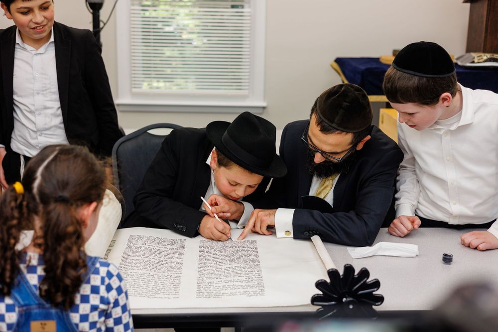 Jewish boys in formal attire studying Torah scroll together at a table during a religious ceremony.