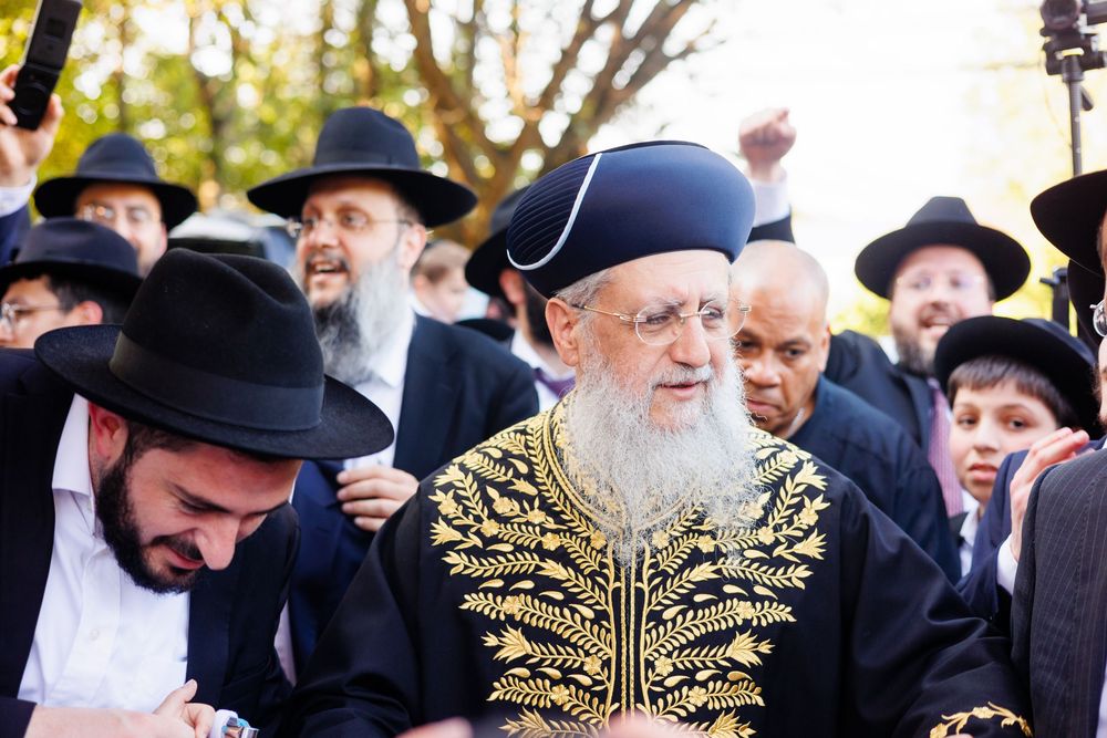 Elderly rabbi in ornate black and gold robe surrounded by crowd of men in traditional Jewish attire outdoors.
