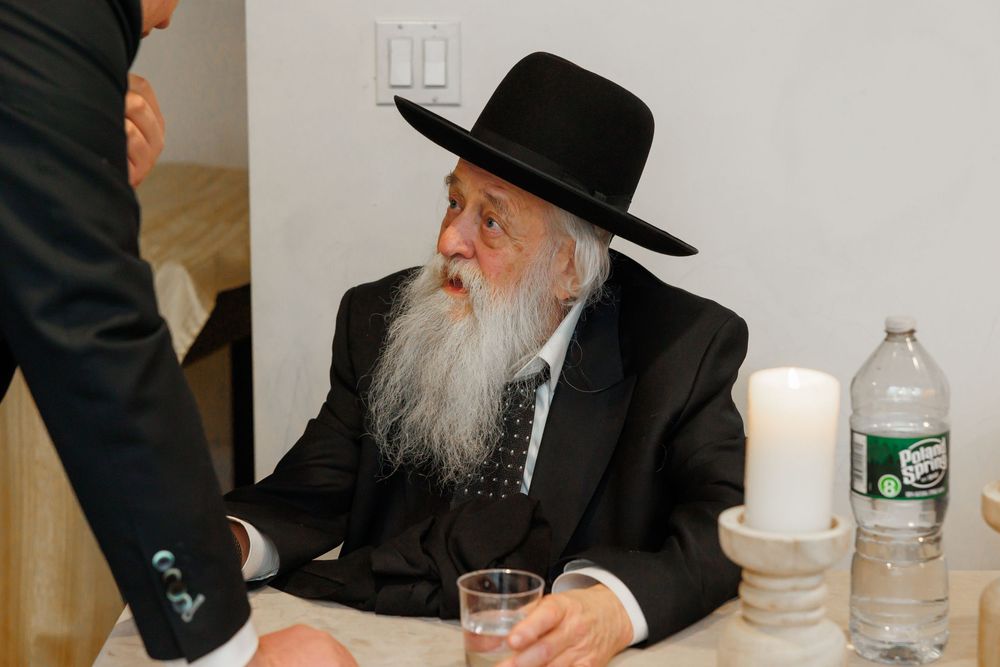 Elderly Jewish rabbi with long white beard wearing traditional black hat and suit seated at a table.