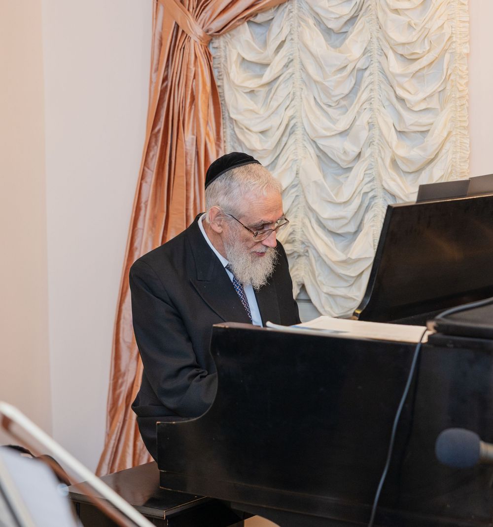 Elderly Jewish man with white beard wearing kippah playing grand piano at formal event.