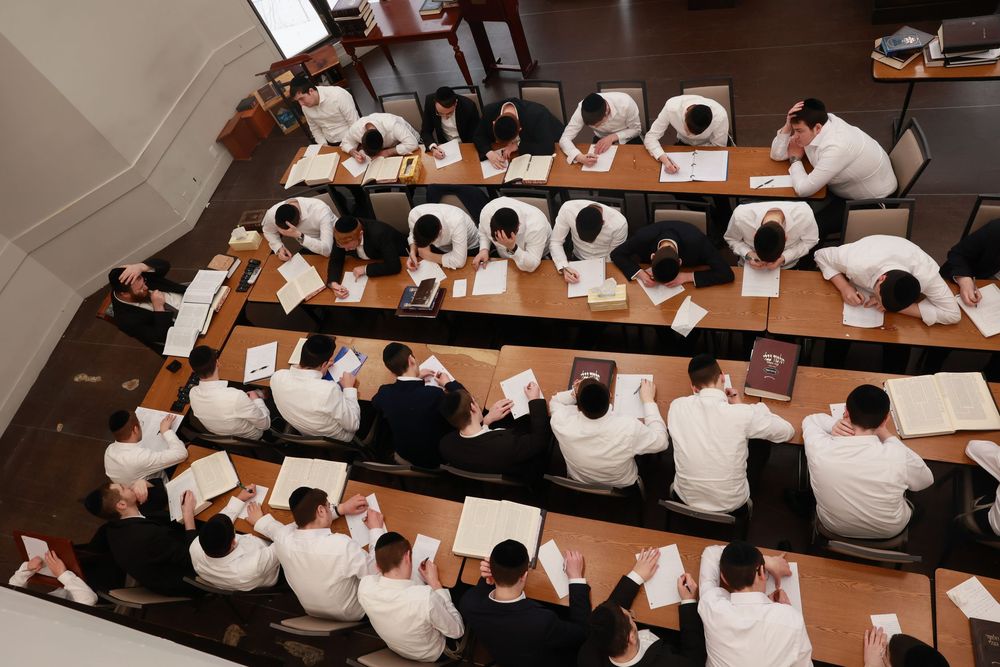Aerial view of students in white shirts studying religious texts at long wooden tables in a yeshiva classroom.