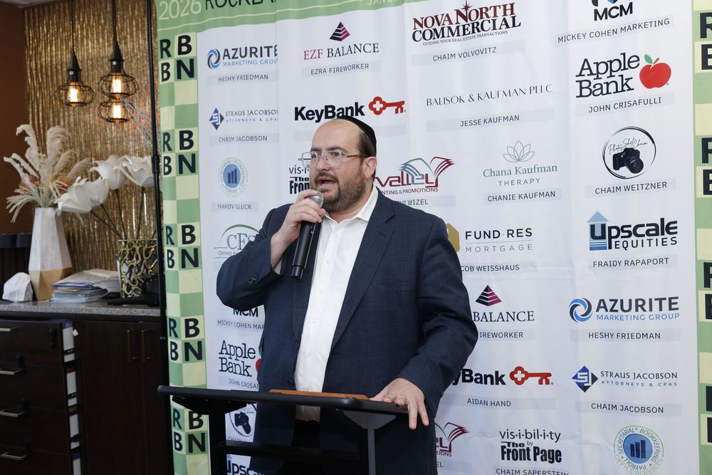 Man in suit speaking at podium with microphone at business networking event with sponsor banners.