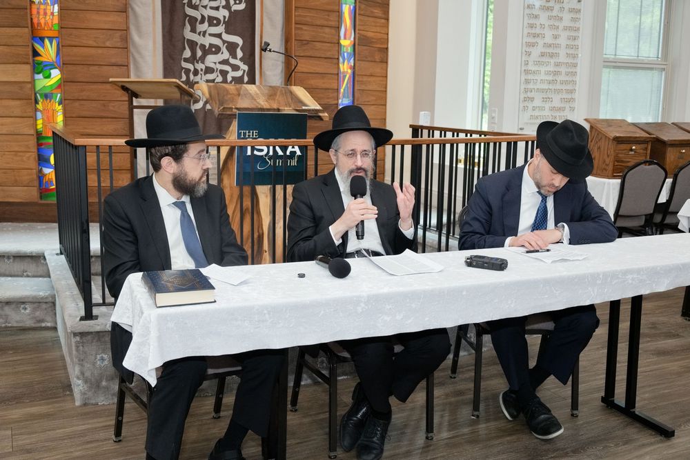 Three Jewish men in traditional attire sit at a white table during a panel discussion at an ISIVA event.