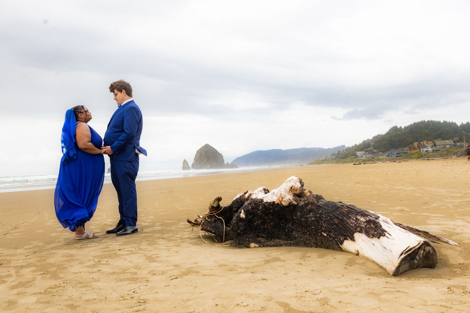 Couple in blue attire poses romantically on Cannon Beach sand near Haystack Rock with a washed-up whale carcass nearby.