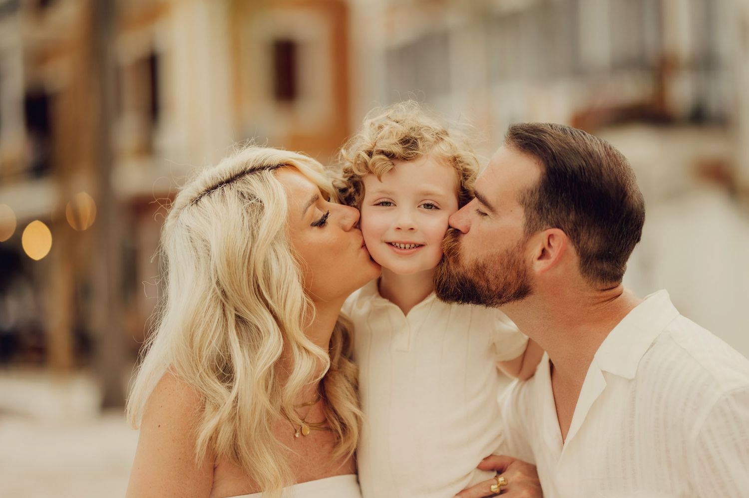 A couple in white wedding attire kisses their smiling curly-haired child on the cheeks outdoors.