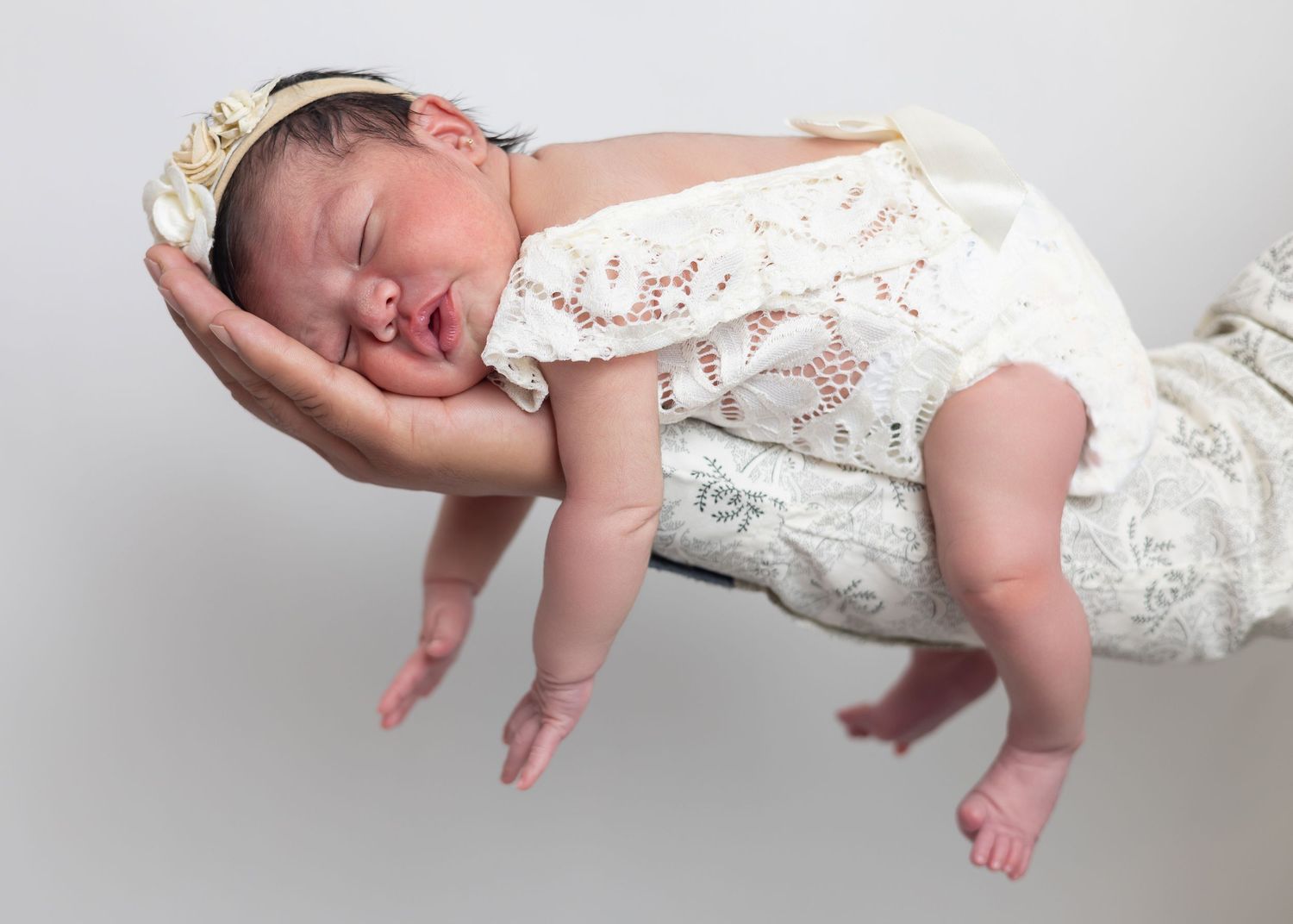Newborn baby in white lace outfit sleeping peacefully on an adult hand against a gray background.
