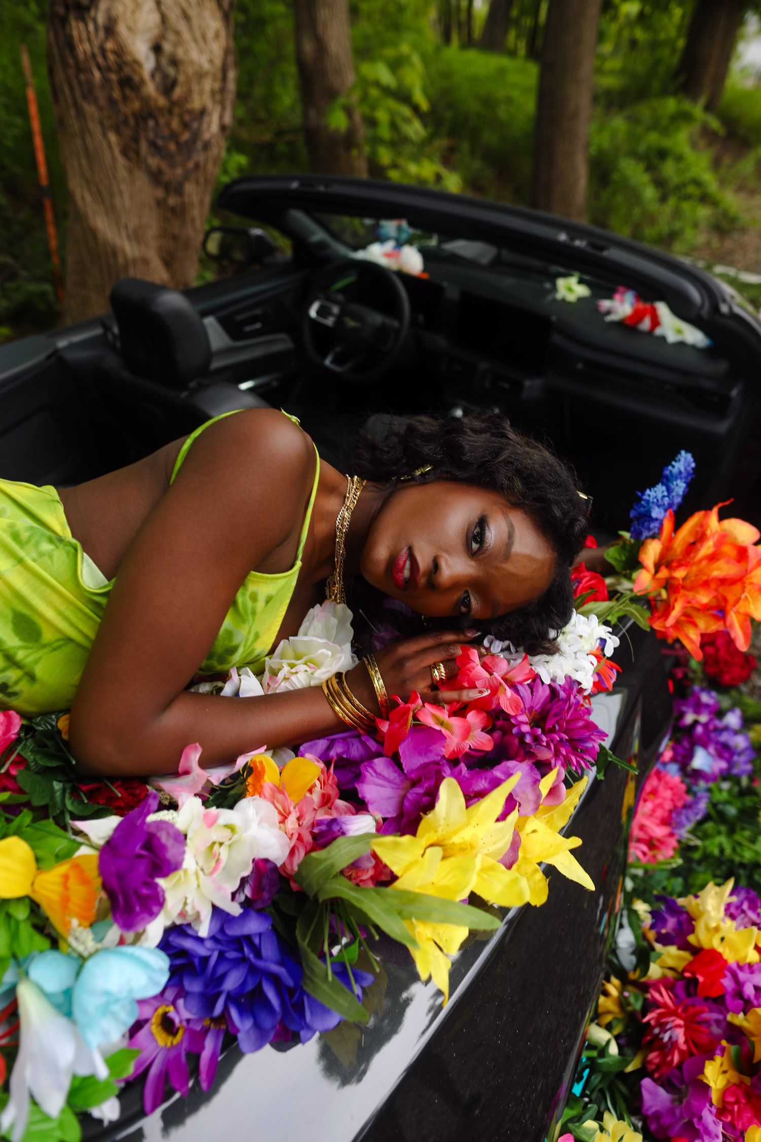 Woman in green dress adorned with colorful flowers reclines in convertible car surrounded by blooms.