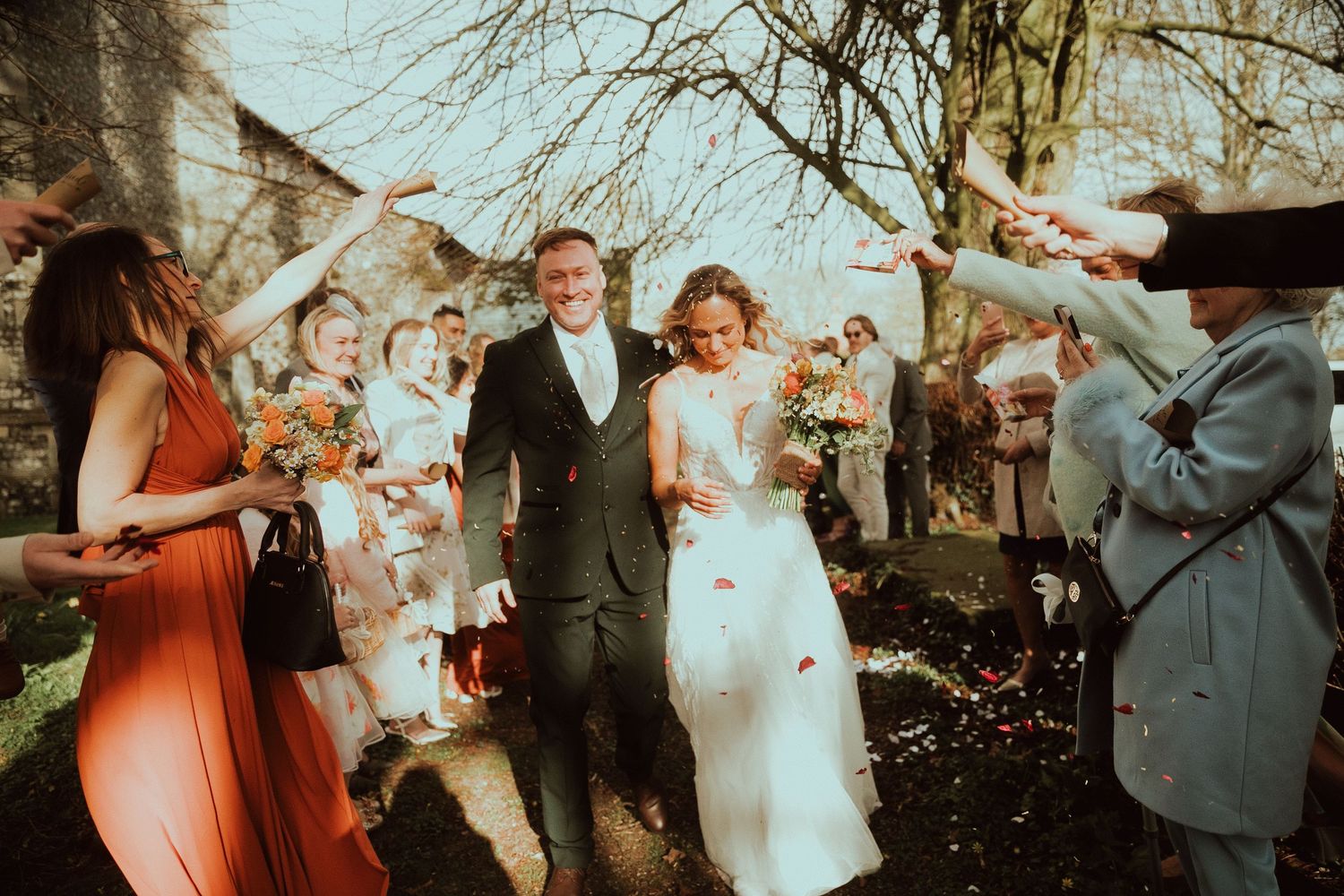 Bride and groom walk through confetti shower surrounded by cheering wedding guests outdoors.