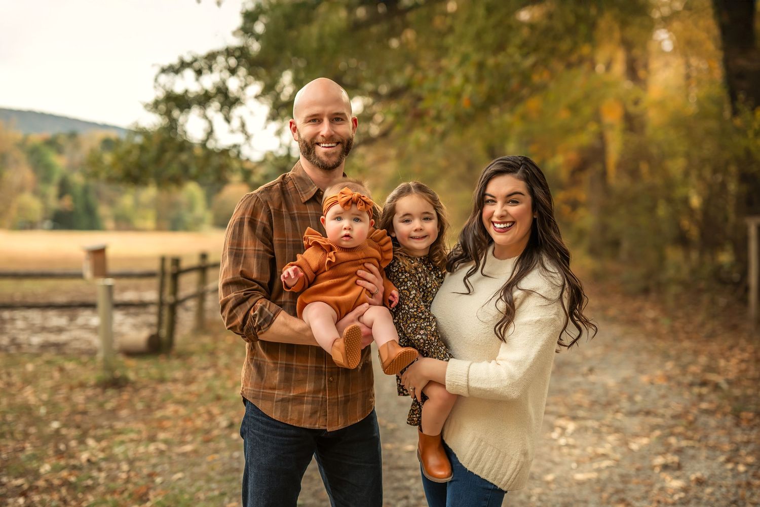 Family of four posing outdoors in fall foliage, parents holding two young children on a scenic country path.