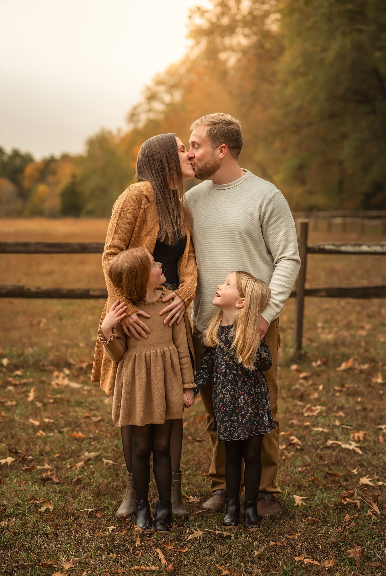 Family of four poses outdoors in autumn, parents kissing while two blonde daughters look up at them near a wooden fence.