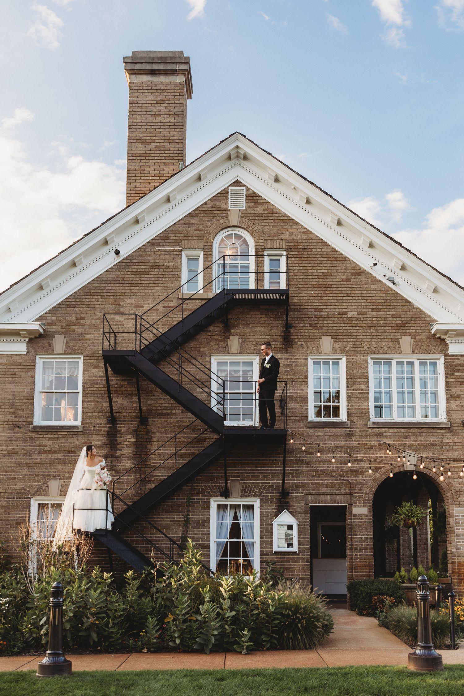 Bride and groom pose on exterior staircase of elegant brick wedding venue with string lights and blue sky.