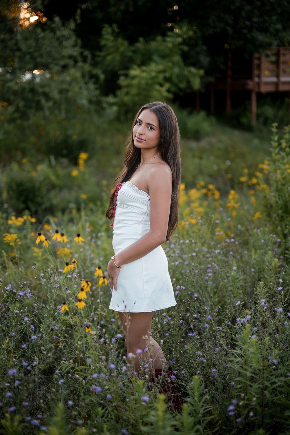 Young woman in white strapless dress standing in wildflower meadow with yellow and purple blooms at golden hour.