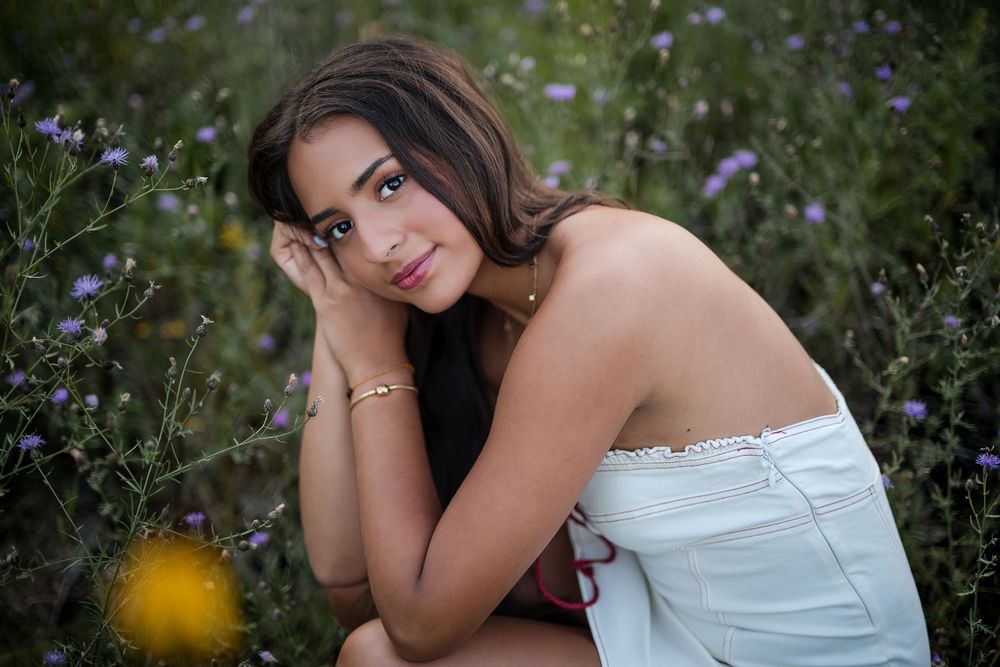 Young woman in white off-shoulder top poses among purple wildflowers in a lush garden setting.