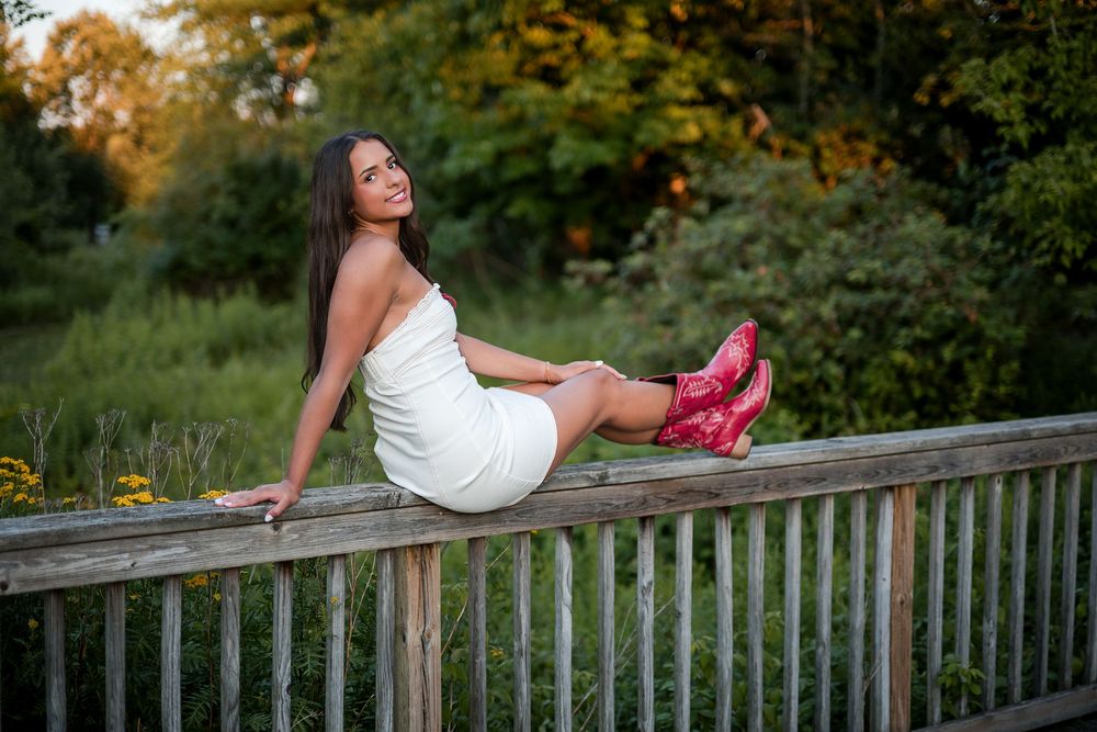 Woman in white dress and red boots sitting on wooden bridge railing surrounded by lush green trees.