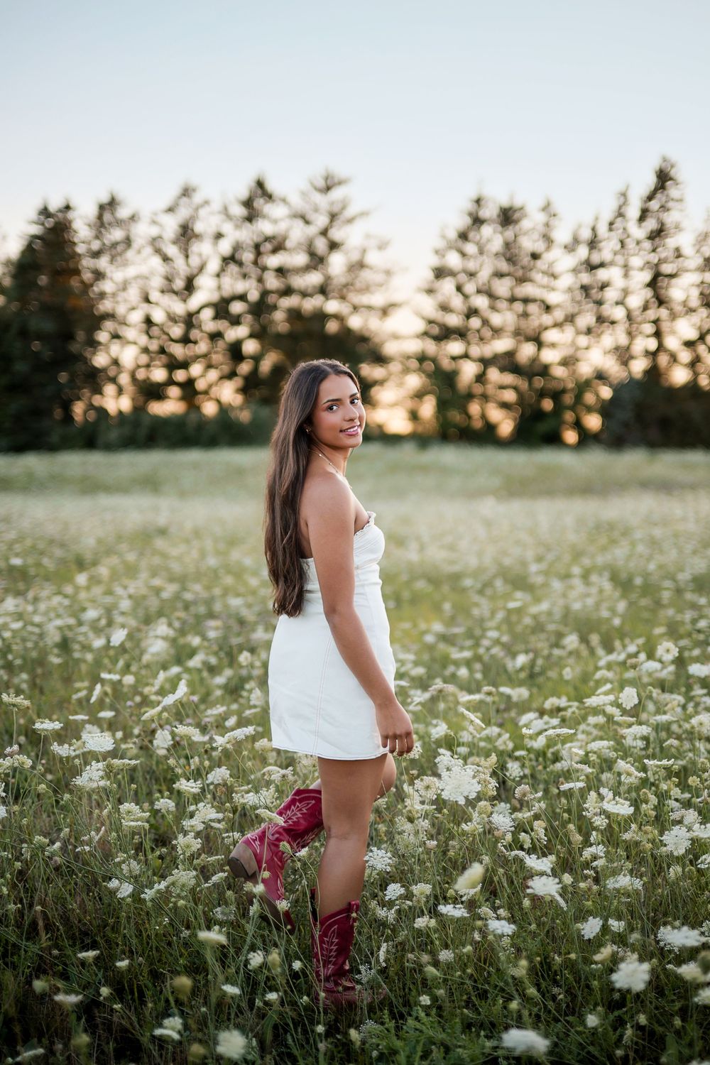 Young woman in white dress and red cowboy boots standing in a wildflower field at golden hour.