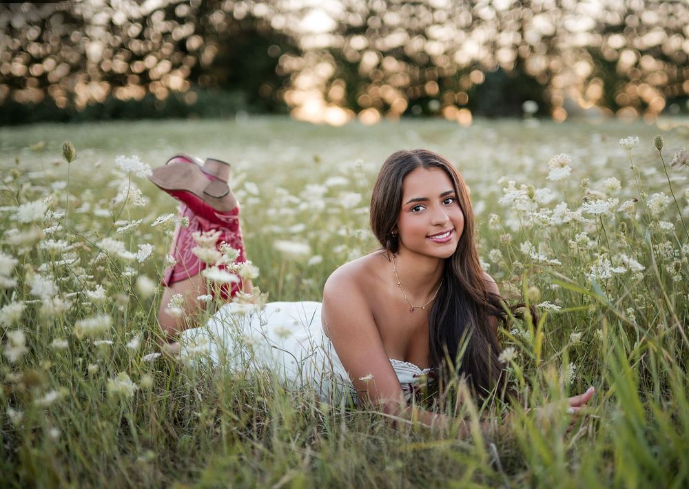 Young woman in white dress lying in a wildflower meadow at golden hour, smiling warmly at camera.