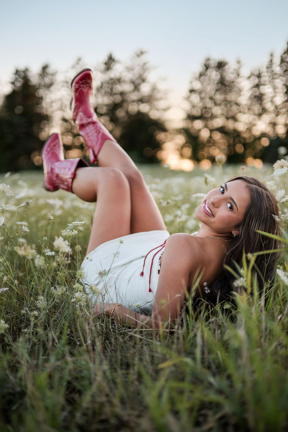 Young woman in white dress and red cowboy boots lying in a wildflower field, smiling at sunset.