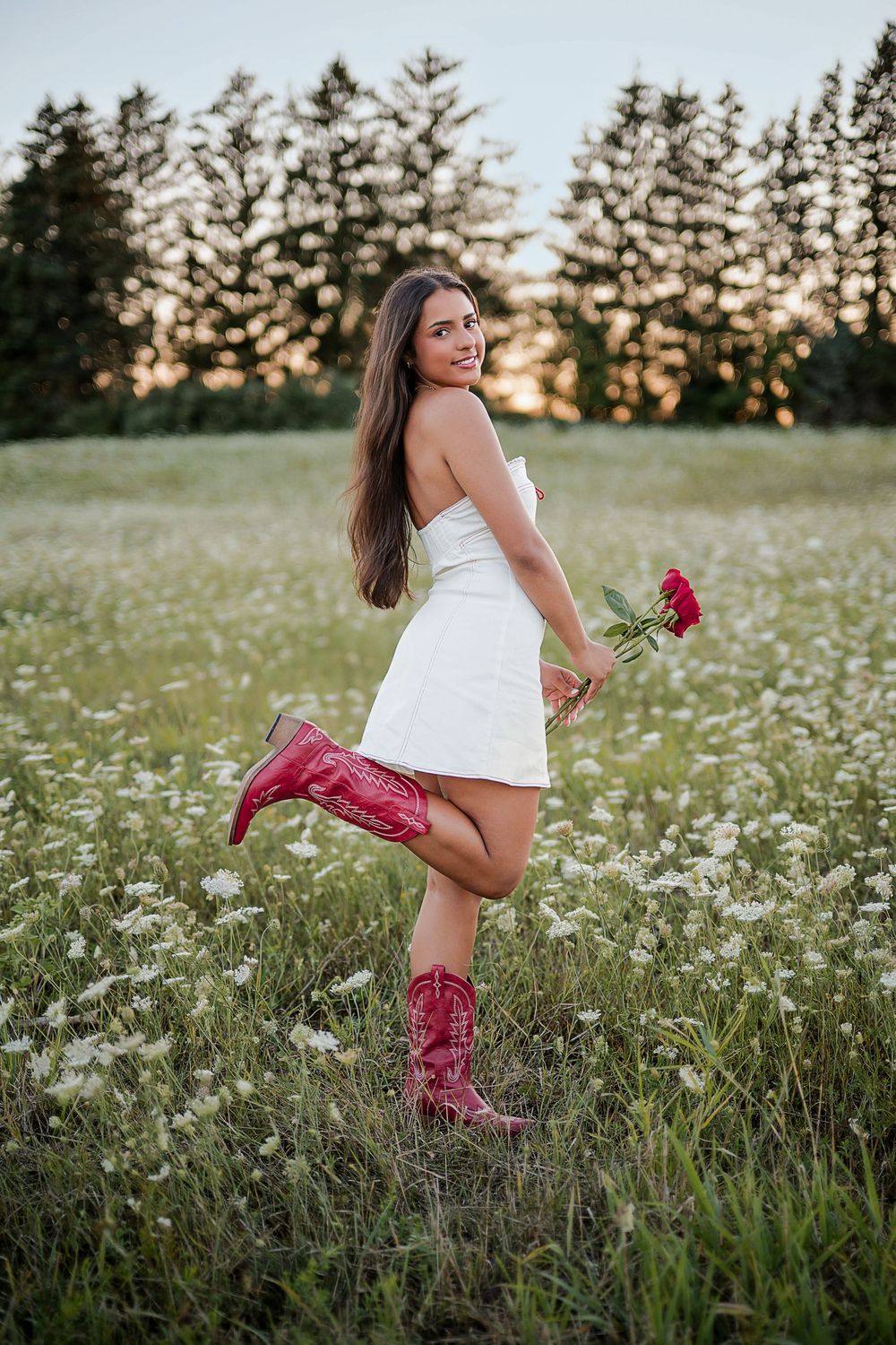 Woman in white dress and red cowboy boots holding roses in a wildflower field at sunset.