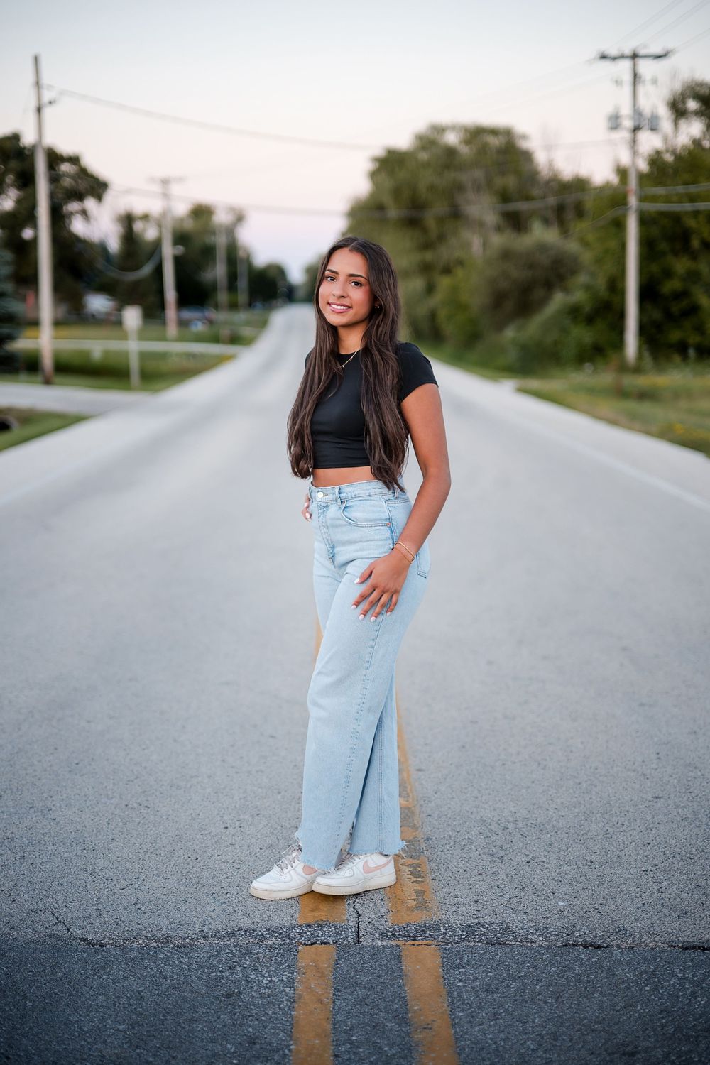 Young woman in black crop top and light blue wide-leg jeans standing on a rural road, smiling confidently.