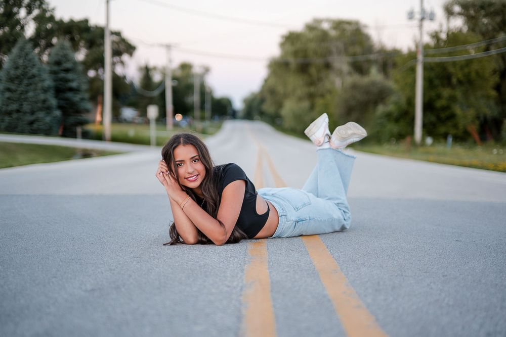 Young woman in jeans and black crop top lying on a rural road centerline, smiling at camera.