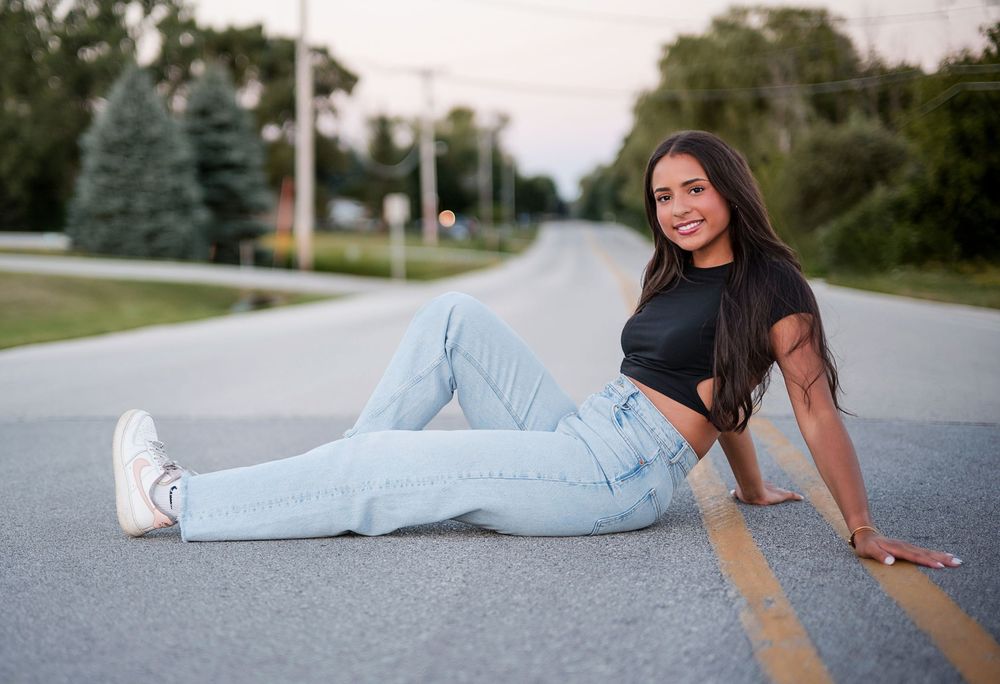 Young woman in jeans and black crop top sitting on a road, smiling confidently with trees in the background.