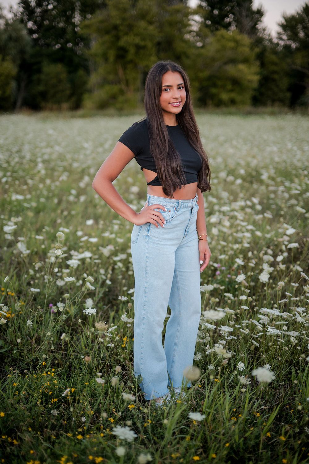 Young woman in black crop top and wide-leg jeans posing in a field of white wildflowers.