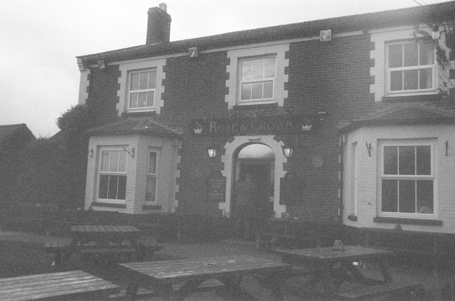 Black and white photo of a traditional brick British pub with arched doorway, sash windows, and outdoor picnic benches.