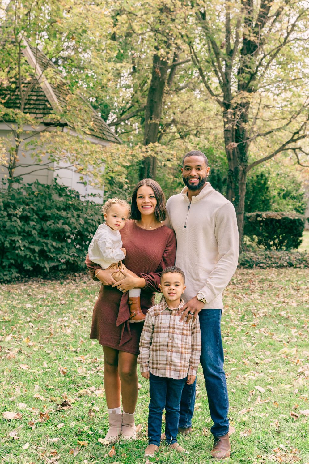 Family of four posing outdoors in autumn, parents with two young children smiling among fall foliage and green grass.