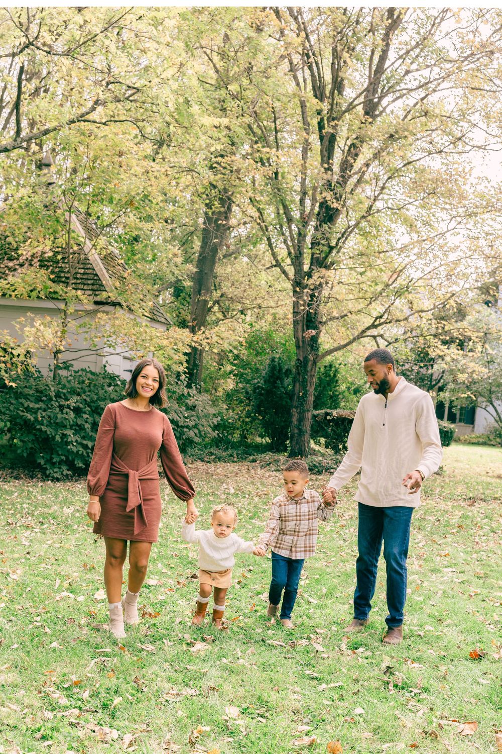 Family of four walking outdoors in autumn, parents holding children's hands on green grass with fall foliage trees.