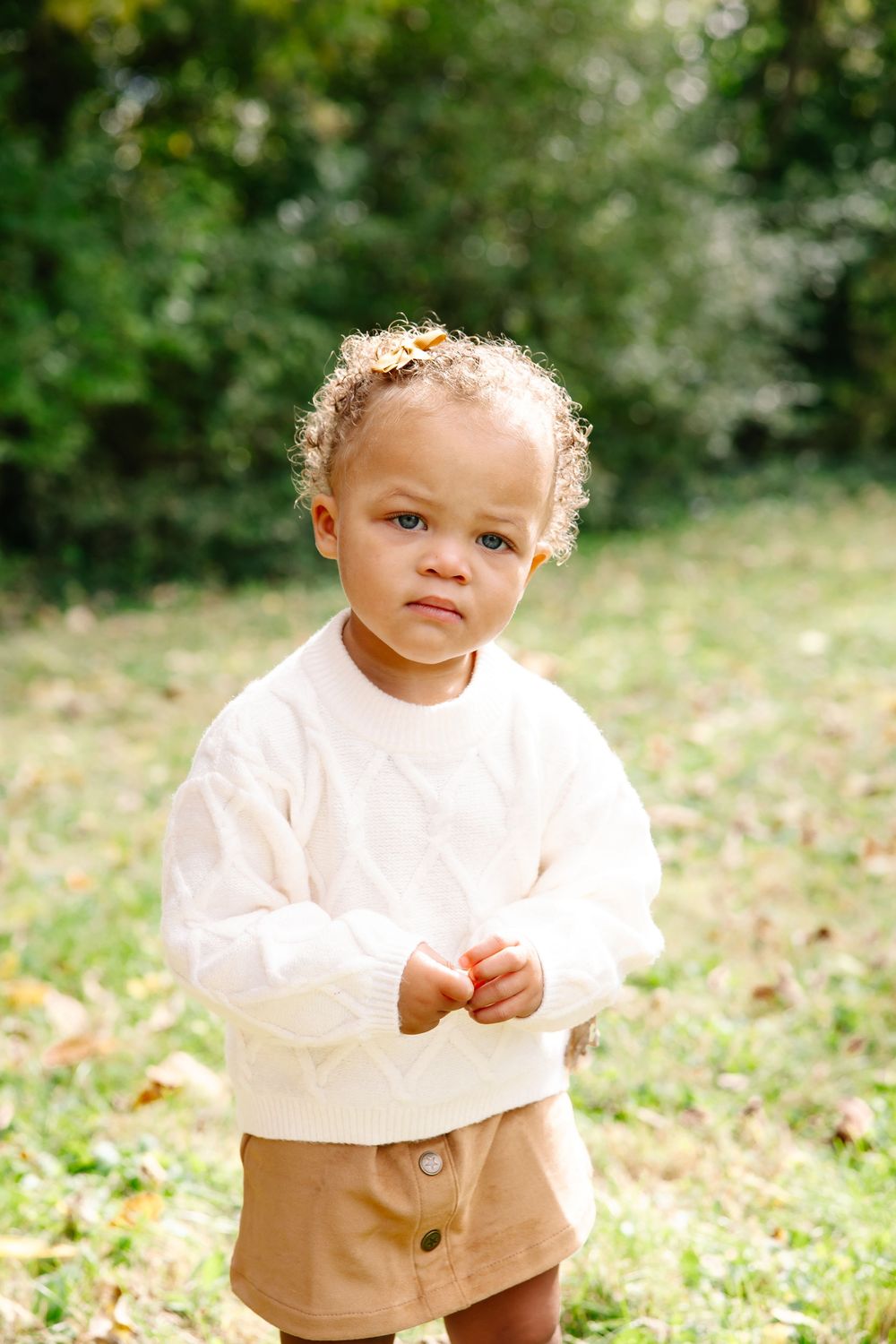 Toddler in white knit sweater and tan shorts standing on grass in a park with green trees in background.