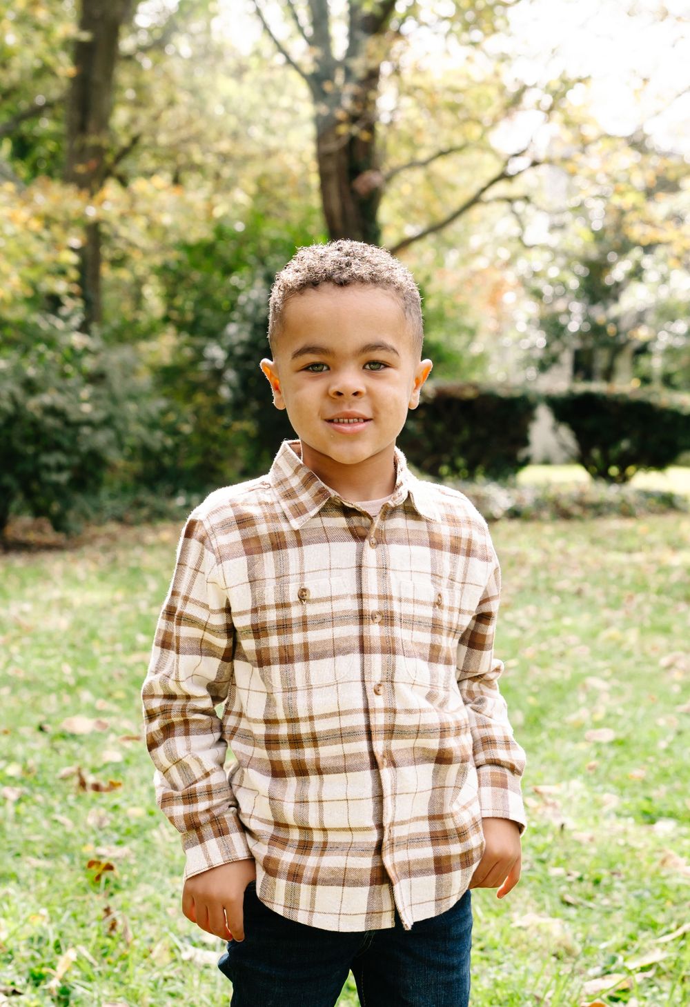 Young boy in plaid shirt smiling outdoors in a lush green park during autumn.
