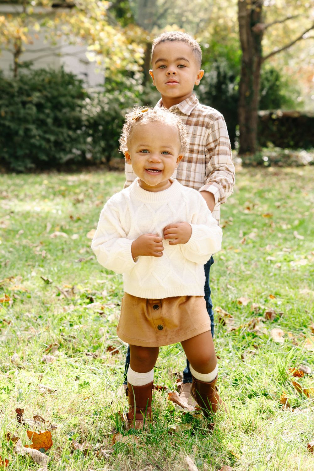 Two young children pose outdoors in autumn, wearing coordinating neutral tones, surrounded by green grass and fallen leaves.