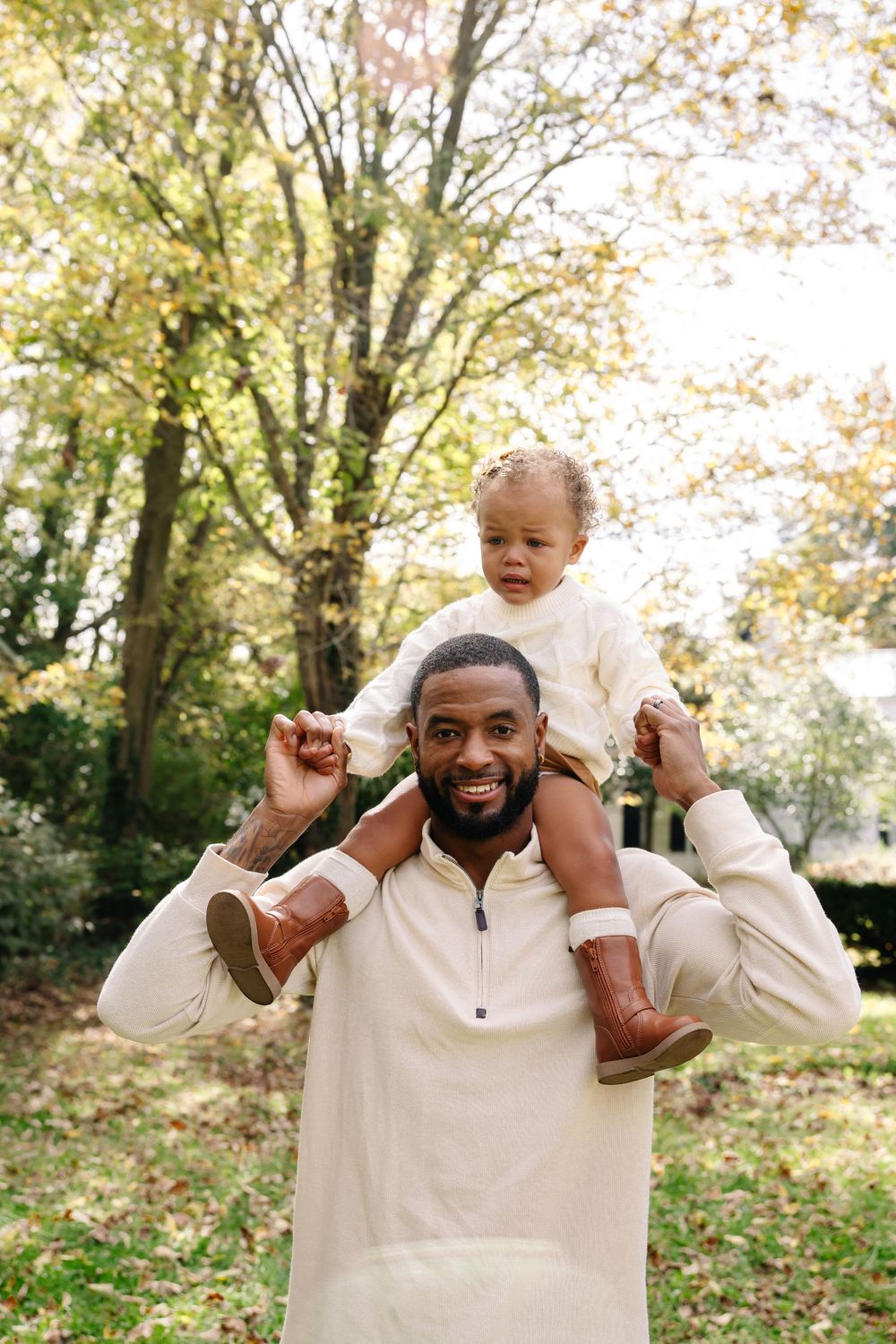 A smiling Black father carries his toddler on his shoulders outdoors among autumn trees, both wearing cream outfits.