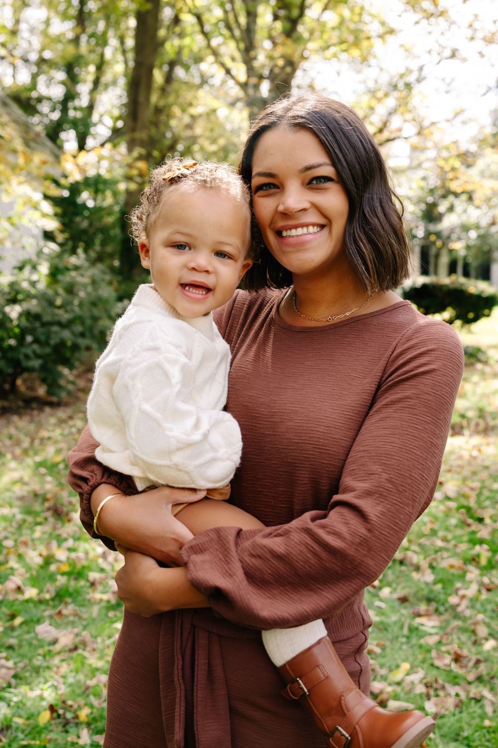 Smiling woman in brown dress holding a happy toddler in white outfit outdoors in a lush green park setting.